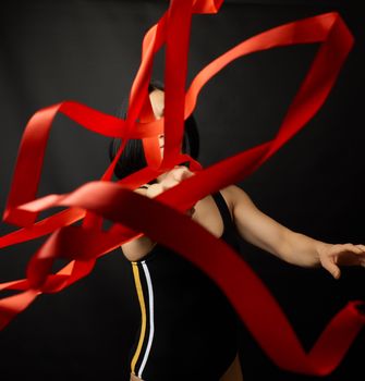 young woman gymnast of Caucasian appearance with black hair spins red satin ribbons, gymnastic exercises on black background, selective focus