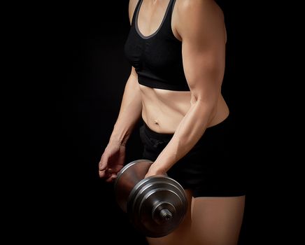 slender girl with a sports figure and muscles holds a type-setting steel dumbbell, she is dressed in a black bra and short shorts, sports backdrop 