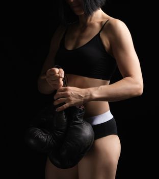 young girl with black hairs and a muscular body holds a pair of old black boxing gloves, she is dressed in a black bra and shorts, low key