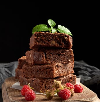 stack of baked square pieces of chocolate brownie cake on brown wooden cutting board, black background