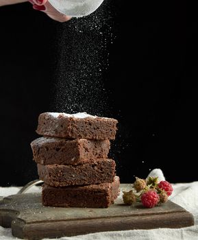 stack of square baked brownie chocolate cake slices sprinkled with white sugar from an iron sieve, small particles fly down, close up