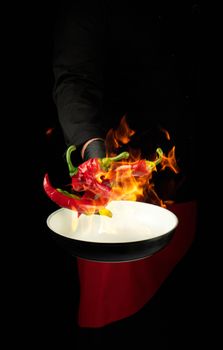 chef in a black uniform holds a round pan and throws up red and green whole chili peppers in a burning fire, cooking spices, low key, copy space