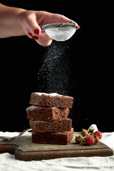 stack of square baked brownie chocolate cake slices sprinkled with white sugar from an iron sieve, small particles fly down, low key