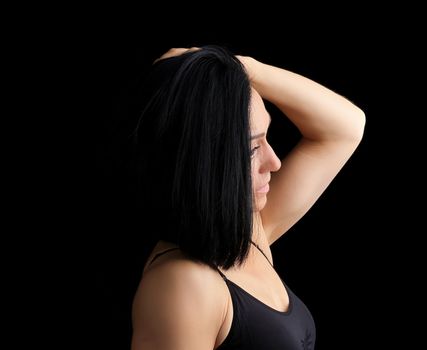 Adult girl with a sports figure in black bra standing on a dark background, model is turned sideways and straightens her black hair, low key