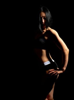 Adult girl with a sports figure in black bra and black shorts standing on a dark background, muscular body, black hair