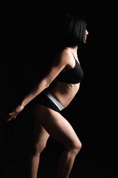beautiful athletic woman with a muscular body is dressed with a black topic and shorts stands sideways, leg is set forward and the arms are extended backwards, studio photo in low key