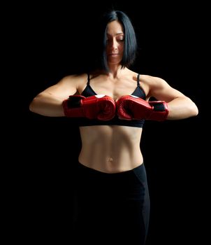 beautiful brunette sporty-looking girl in a black bra and leggings stands on a dark background, wearing red boxing gloves on her hands