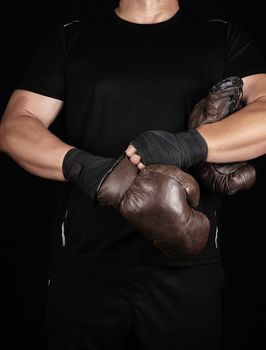 adult muscular man in black clothes puts on leather brown boxing gloves on his hands before a competition, his hands are wrapped in a black sports bandage