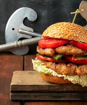 homemade double hamburger with pork fried steak, red tomatoes, fresh round bun with sesame seeds on a vintage wooden board