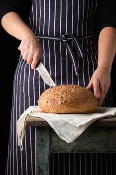 woman in an blue apron with a knife in her hand about to cut round baked bread on a wooden table, dark background