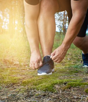 athlete in black uniform crouched and tying the laces on blue sneakers in the middle of the forest, concept of a healthy lifestyle and jogging