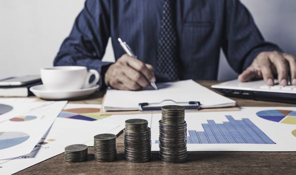 Business man working and writing on notebook with stack of coins for financial and accounting concept.