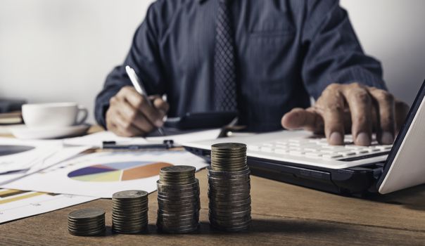 Business man working and writing on notebook with stack of coins for financial and accounting concept.