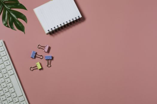 Top view with the keyboard and notebook on pink background. Cluttered office desk background.