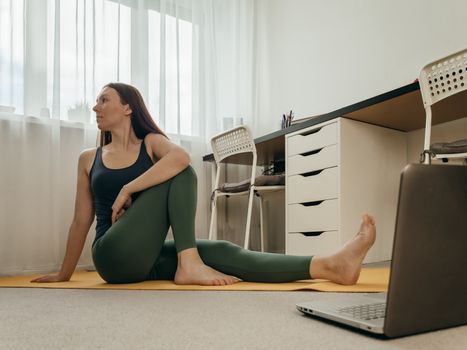 Young woman sitting on floor and doing yoga or stretching at home near laptop. Yoga online in small space home and wellbeing concept. Copy space for text or design.