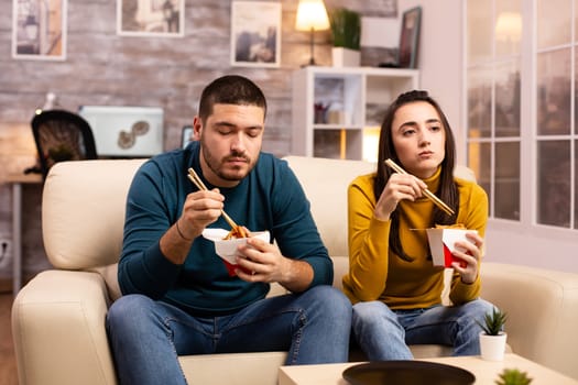 In modern cozy living room couple is enjoying takeaway noodles while watching TV comfortably on the sofa