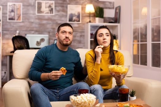 Beautiful young couple watching TV and eating fast food takeaway in the living room