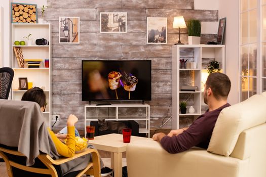 Back view of couple in living room watching a movie on the TV while eating takeaway food