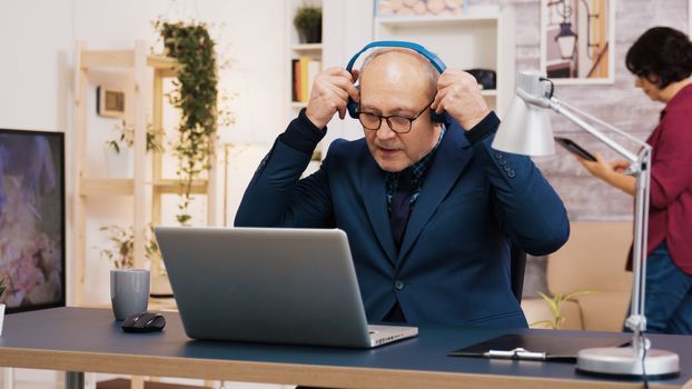Senior old man listening music on headphones while working on laptop in living room. Wife using tablet in the background.