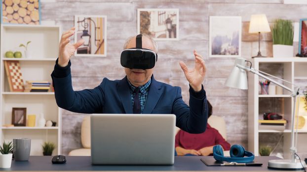 Senior man using virtual reality goggles in living room. Woman watching tv while sitting on the sofa in the background.