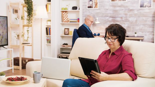 Zoom in shot of elderly age woman using tablet sitting on coich. Senior man reading a book in the background.