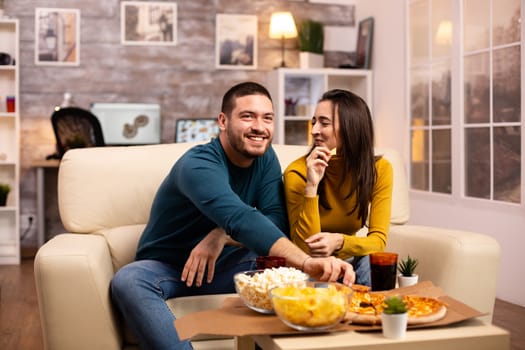 Beautiful young couple watching TV and eating fast food takeaway in the living room