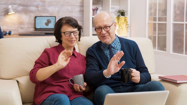 Elderly age couple wave at laptop during a video call with their family.