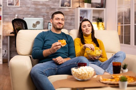 Beautiful young couple watching TV and eating fast food takeaway in the living room