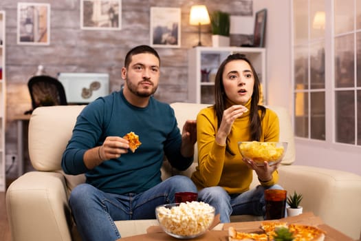 Beautiful young couple watching TV and eating fast food takeaway in the living room