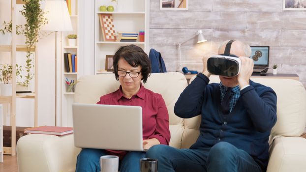 Senior man trying a VR headset in the living room while his wife uses a laptop next to him. Modern old couple using technology