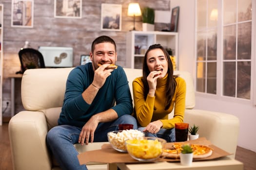 Beautiful young couple watching TV and eating fast food takeaway in the living room