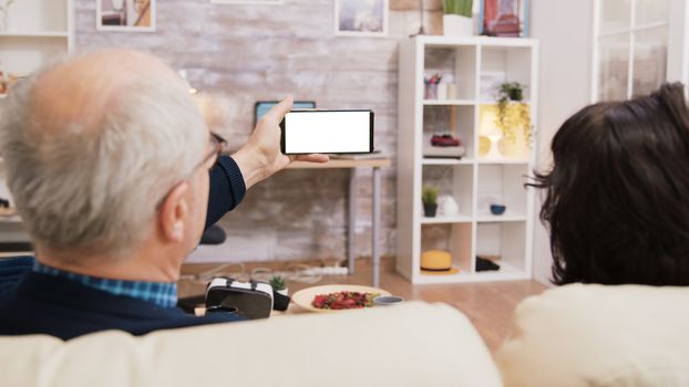 Back view of elderly age couple looking at phone with green screen while sitting on sofa in living room.