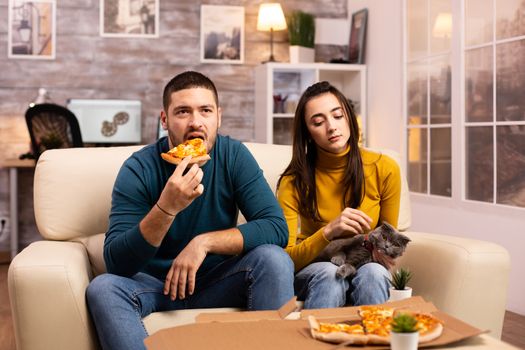 Gorgeous young couple eating pizza while watching TV in the living room sitting on the sofa