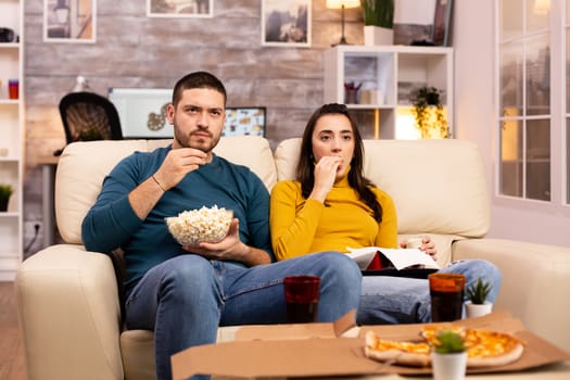 Beautiful young couple watching TV and eating fast food takeaway in the living room