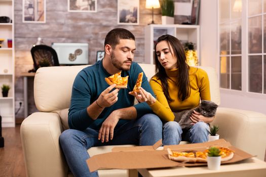 Gorgeous young couple eating pizza while watching TV in the living room sitting on the sofa