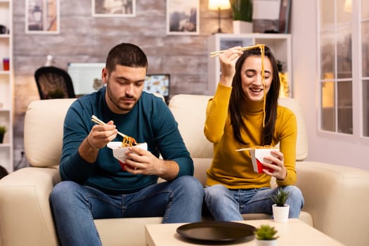 In modern cozy living room couple is enjoying takeaway noodles while watching TV comfortably on the sofa