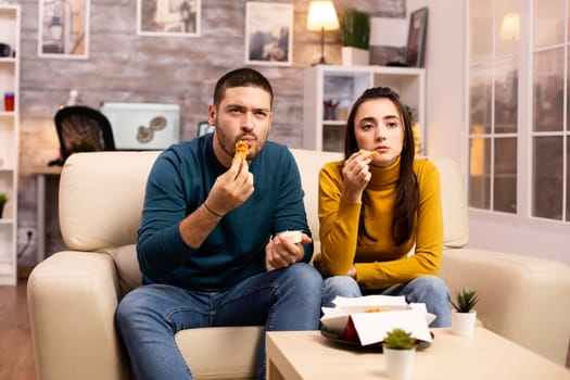 Young couple eating fried chicken in front of the TV in the living room