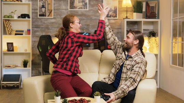 Couple celebrating victory while playing video games using wireless controllers and giving high five.