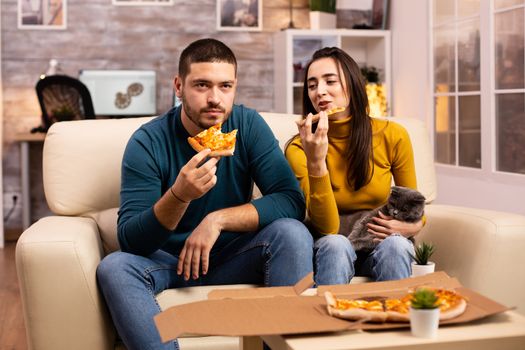 Gorgeous young couple eating pizza while watching TV in the living room sitting on the sofa