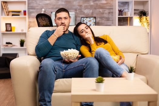 Handsome couple at home eating pop corn and watching TV on the sofa in the living room