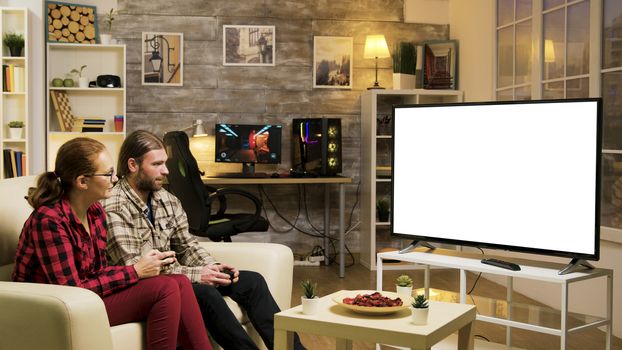 Cheerful couple sitting on couch playing video games on tv with green screen using wireless controllers in living room.