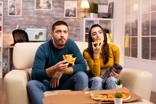 Gorgeous young couple eating pizza while watching TV in the living room sitting on the sofa