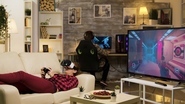 Woman lying on sofa playing video games using vr headset in living room. Boyfriend playing on computer.