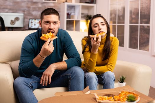 Gorgeous young couple eating pizza while watching TV in the living room sitting on the sofa