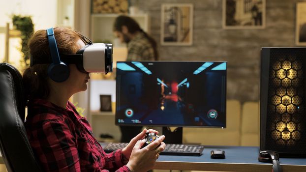 Professional female gamer using vr headset and wireless controller to play computer games. Man texting on phone in the background.