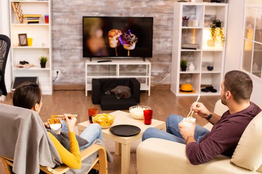 Back view of couple in living room watching a movie on the TV while eating takeaway food