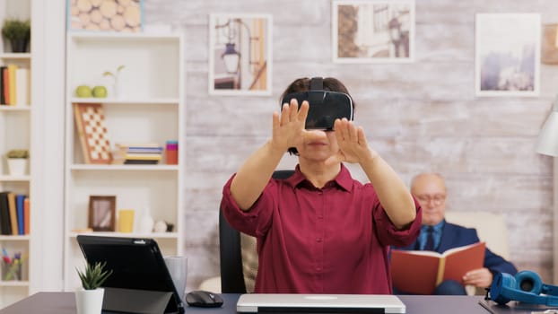 Retired senior woman amazed while using virtual reality goggles in living room. Elderly man sitting on sofa while reading a book.