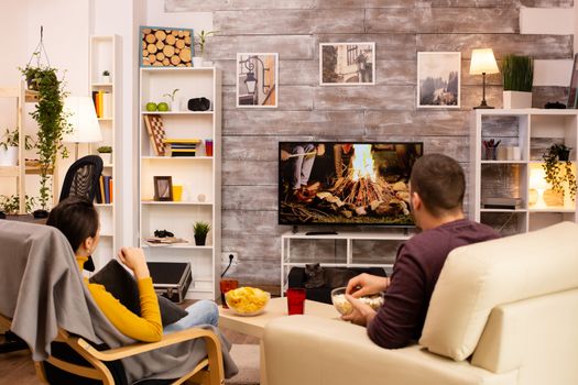 Back view of couple in living room watching a movie on the TV while eating takeaway food