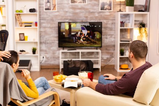 Back view of couple in living room watching a movie on the TV while eating takeaway food