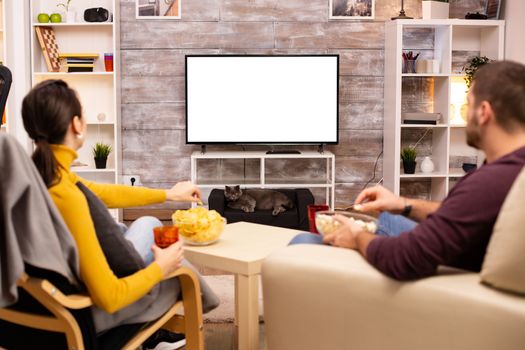 Couple looking at isolated TV screen in cozy living room while eating takeaway food